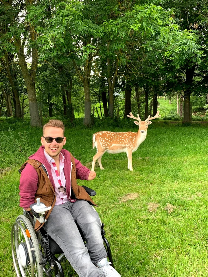 Patrick sitzt im Dubliner Phoenix Park in seinem Rollstuhl und trägt Sonnenbrille. Hinter ihm steht in weniger Entfernung ein Hirsch und schaut ebenfalls in die Kamera.