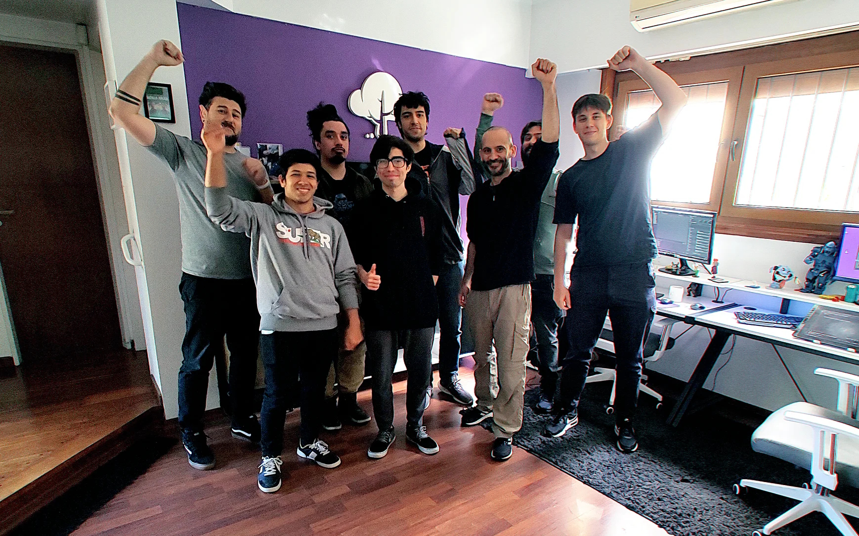 Eight people stand in front of a Purple Tree logo on a purple wall, with their fists pumping in the air.