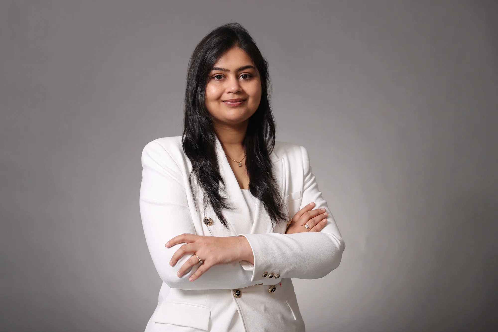 A woman with long black hair wears a white blazer at the camera with her arms folded across her chest.