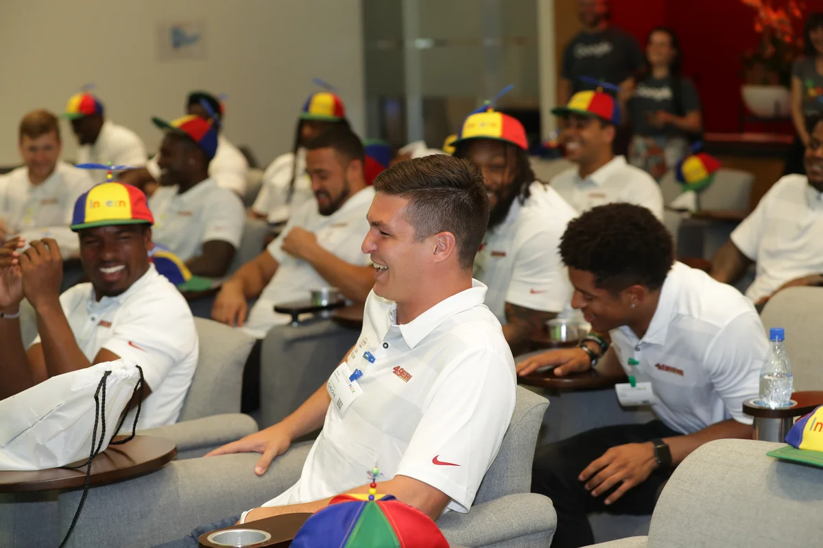 Members of the San Francisco 49ers sitting in chairs wearing intern hats and laughing.