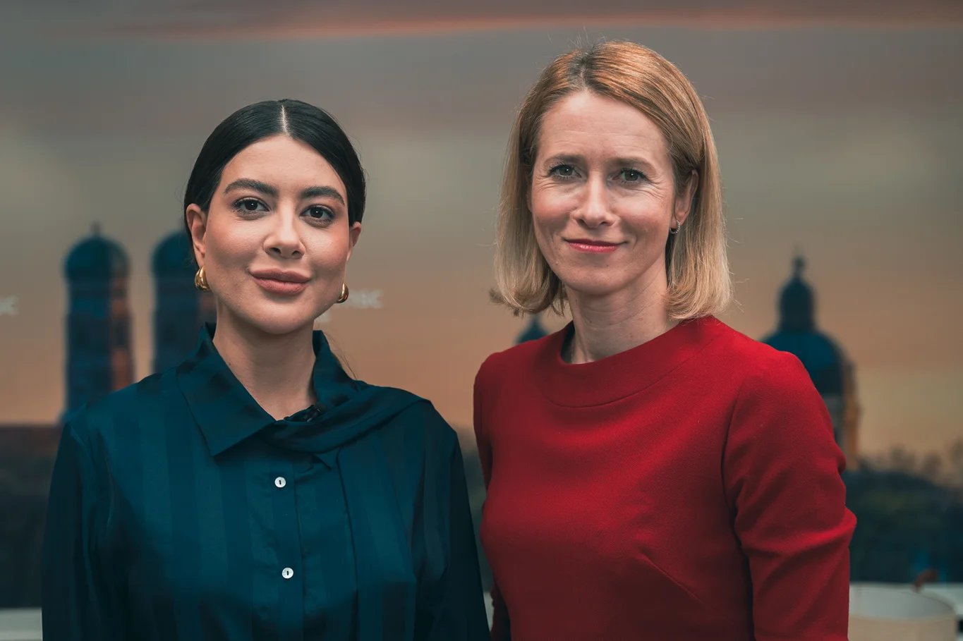 Zwei Frauen lächeln vor der Münchner Skyline, Ramina links mit dunkler Bluse, Kaja Kallas rechts mit rotem Kleid