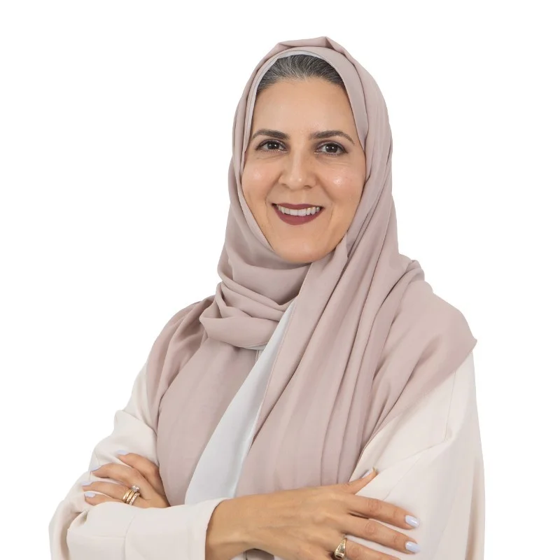 A woman wearing a cream long-sleeve top and light bpink headscarf smiles at the camera, with her arms folded across her chest.