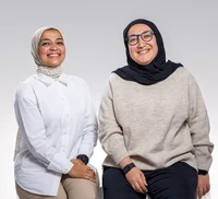 A smiling woman wearing a white shirt and headscarf sits next to a woman wearing a light brown shirt, black glasses and a black headscarf.