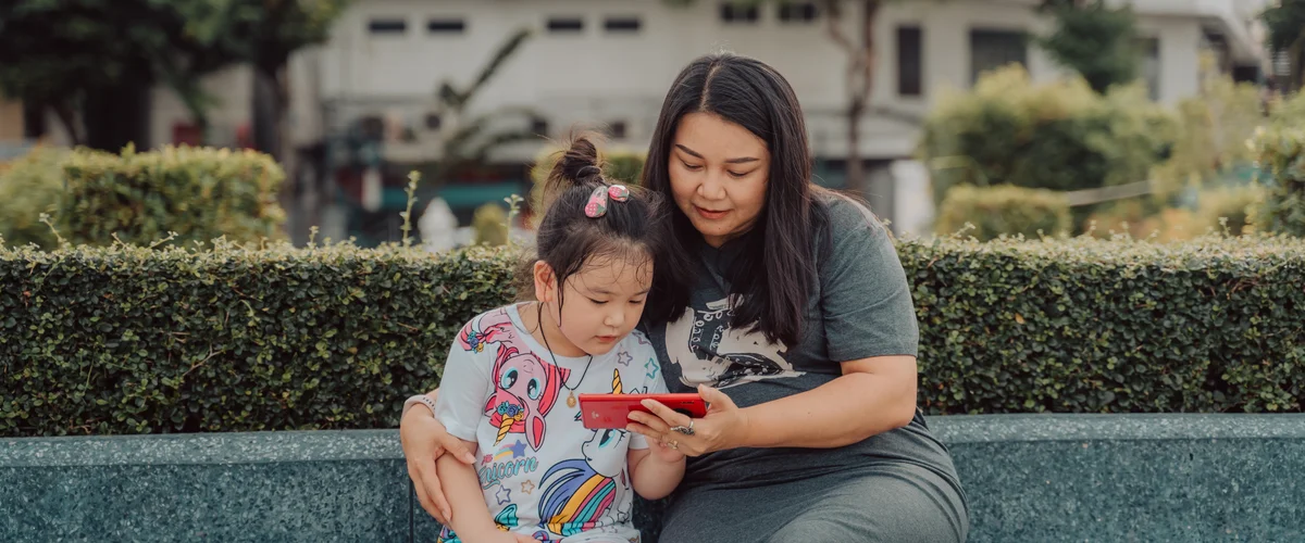 An adult and child sit outside and look at a red Android phone together.