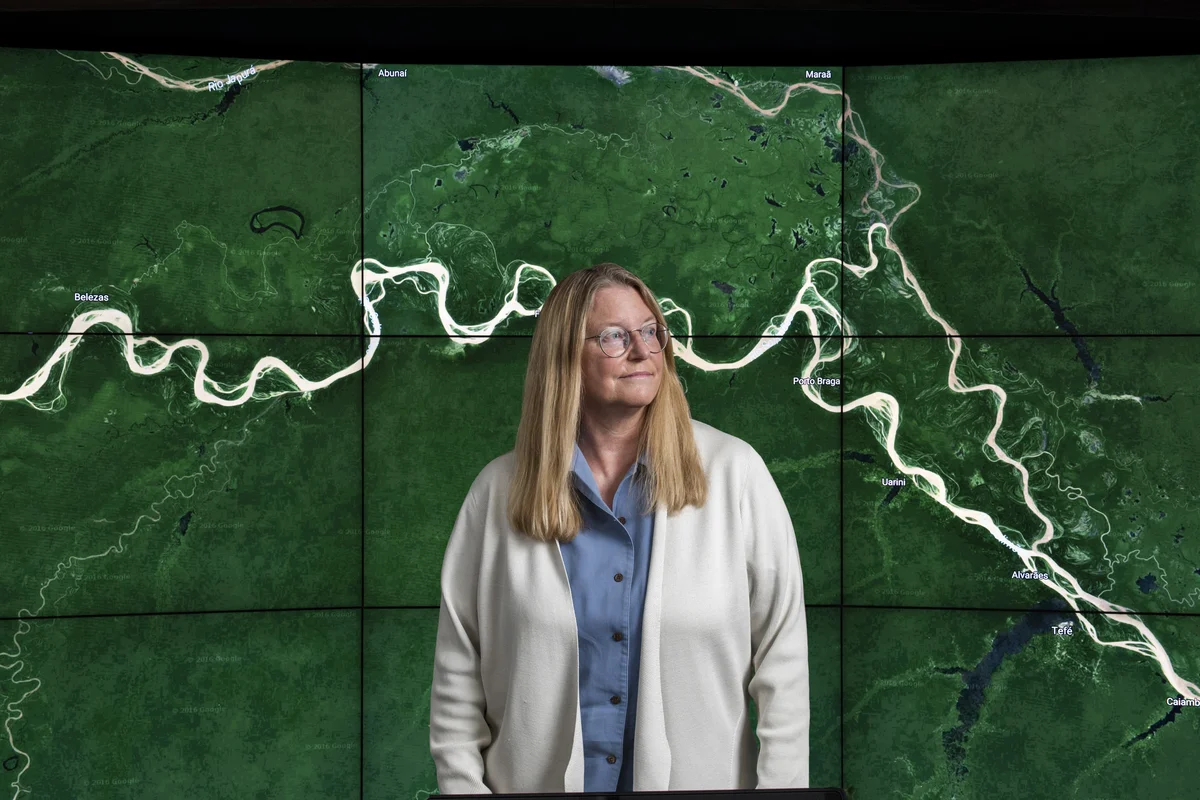 Image of Rebecca Moore in a white cardigan standing in front of a satellite map image.