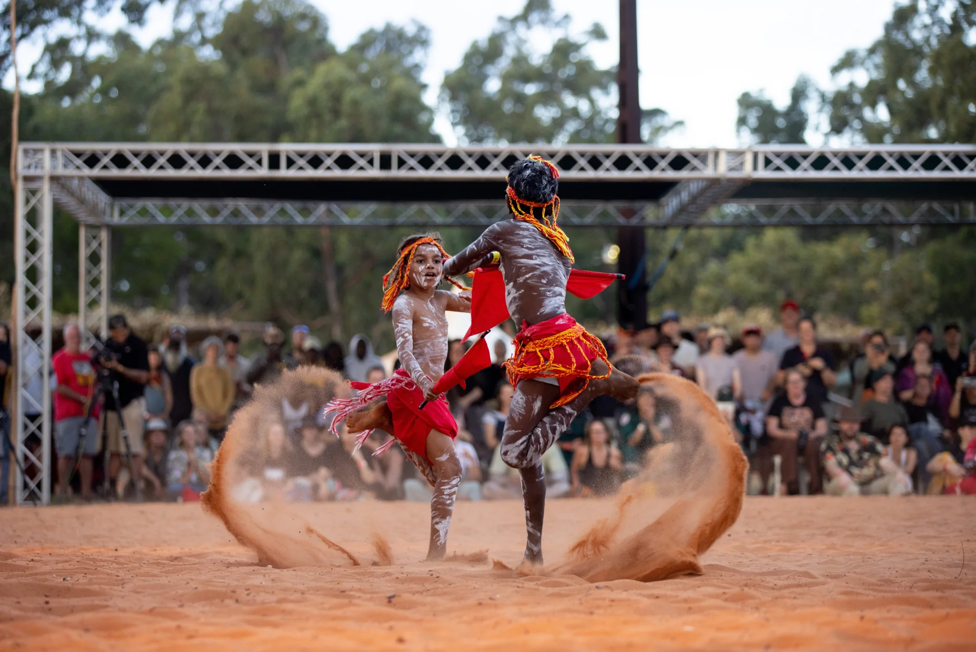 Two boys dance with red flags, and kick-up dirt on Arnhem Land