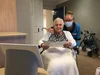 Image shows an elderly woman in a wheelchair with a nurse who is wearing a mask leaning over her shoulder. Bother women are looking at a Google Nest Hub Max and reading a booklet with instructions. The nurse is pointing at the screen, and the woman in the wheelchair is smiling.