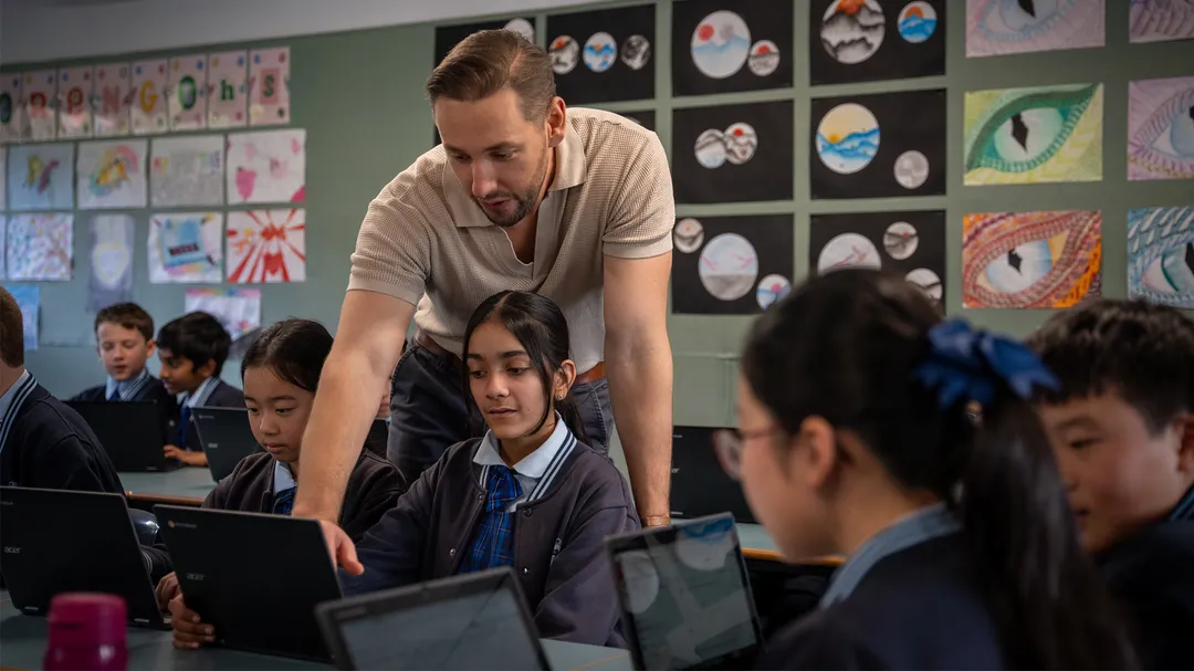 A teacher wearing a tan shirt with light brown hair stands over a student at their laptop computer and points to the screen.