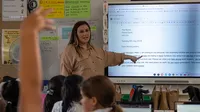 A teacher with dark hair and wearing a tan shirt stands at the front of the room, pointing to a screen while teaching a lesson. A child raises their hand in the back of the classroom.