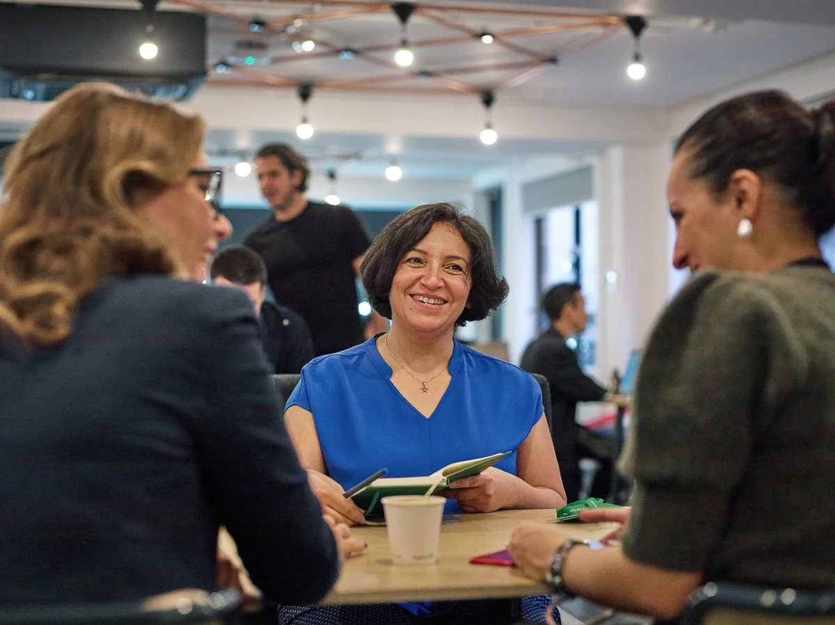 Three women—one in a blue shirt facing the camera, notebook in hand—sit around a coworking desk at a Google for Startups Campus, with other people talking and standing in the background.