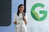 Patricia Florissi, mulher branca de blusa e saia branca, fala com microfone no palco em frente ao logo do Google em verde