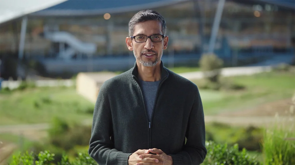 Google CEO Sundar Pichai delivering a speech with Google's Bayview campus and green space in the background. He is wearing a grey zip-up cardigan and blue t-shirt with his hands clasped in front of him.