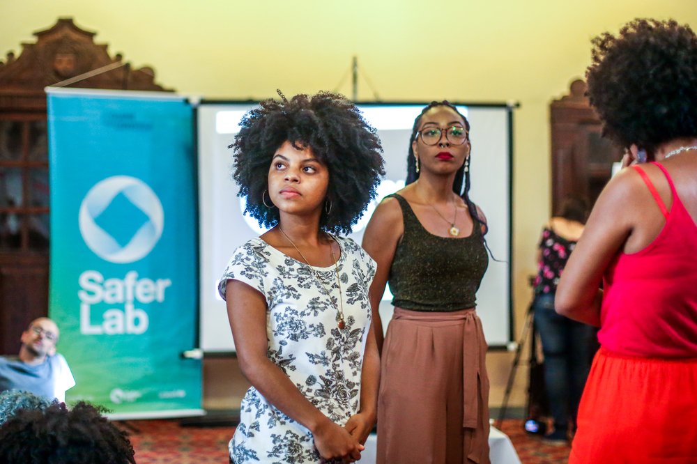 Mulheres negras acompanham event em pé. Painel com símbolo da SaferNet aparece atrás delas, desfocado.