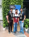 Four men stand in front of a black and blue colored sign that says “Build with AI”. They are all smiling at the camera. In the background are several plants.