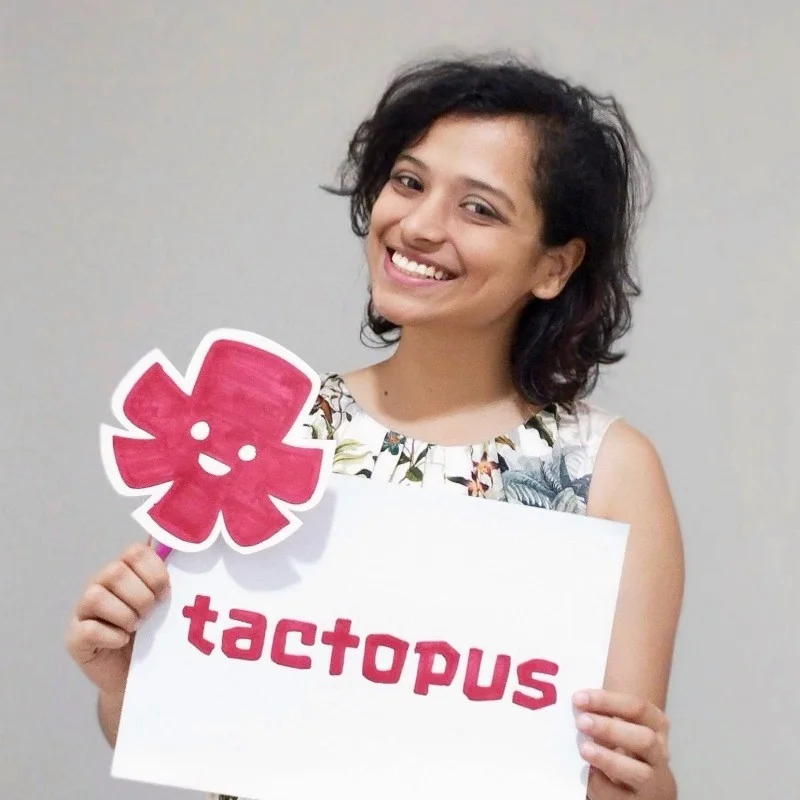 Saloni Mehta, founder of Tactopus Learning Solutions, smiles at the camera with her head tilted slightly to one side. She holds a white sign that reads "tactopus" in pink letters, and wears a sleeveless floral top.