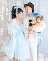 Sam, Kyle, and baby Kiki pose against a shimmery, wintry white backdrop. Sam wears a baby blue princess dress and hair bow, Kyle is in a white suit with a blue scarf and black top hat and baby Kiki wears a yellow dress with a blue pom-pom headband.