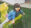 Photo of Sam as a young child. She sits on top of a green bouncy ride outside, wearing a blue dress, white tights and her hair styled with bangs in a bob cut.