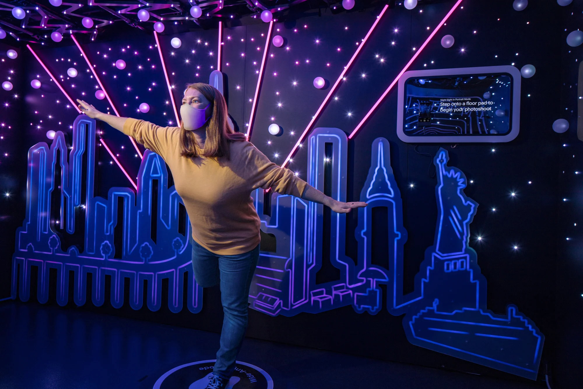 A person poses in front of a neon-lit New York cityscape in a dark room.