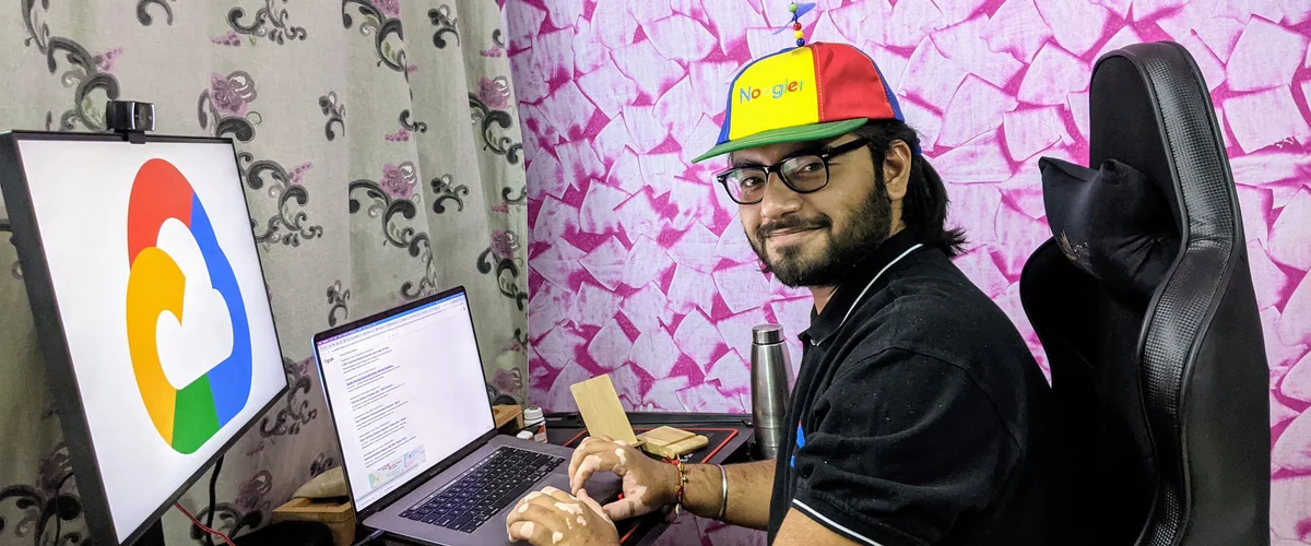 Sanjay sits at his desk at home. He’s wearing a Noogler hat and typing on a laptop. In front of him is a monitor with the Google Cloud logo on the screen.