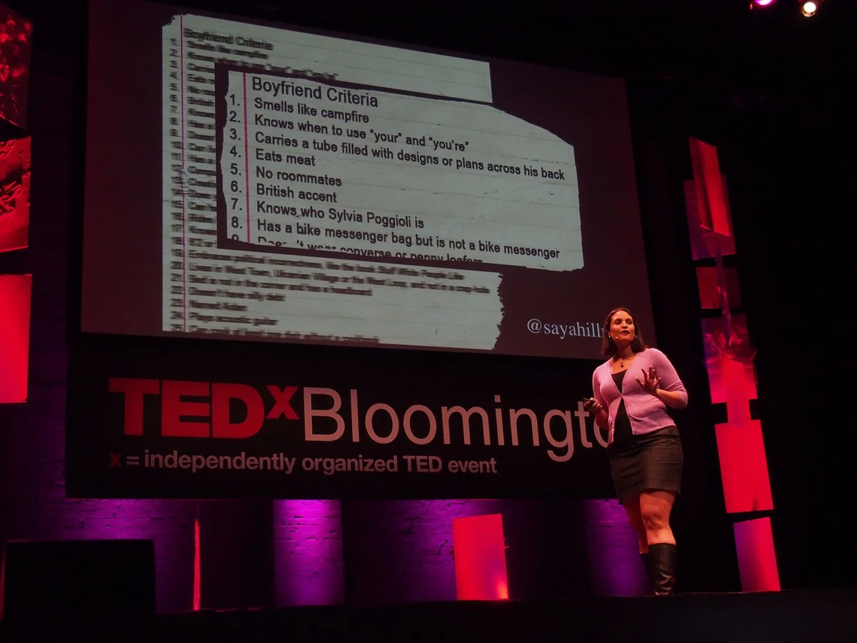 A woman in a pink cardigan, black skirt and black boots speaks on stage at TEDx Bloomington. A large screen behind her shows a detail from a larger list of “Boyfriend Criteria.