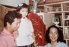 Three people, two adults and one child, are in an old photograph. The child is wearing a white shirt and a white hair pin and looking into the camera. She is leaning on an adult man who is wearing a pink shirt, looking down and smiling. To the left of the child is a woman with dark curly hair, who is looking away from the camera and smiling.