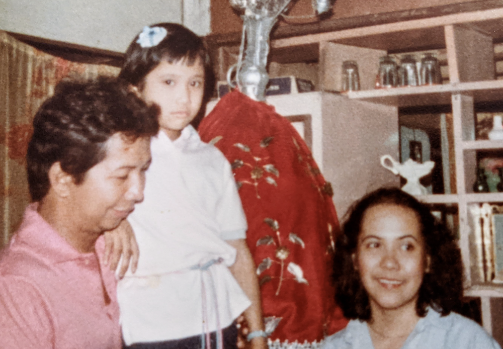 Three people, two adults and one child, are in an old photograph. The child is wearing a white shirt and a white hair pin and looking into the camera. She is leaning on an adult man who is wearing a pink shirt, looking down and smiling. To the left of the child is a woman with dark curly hair, who is looking away from the camera and smiling.