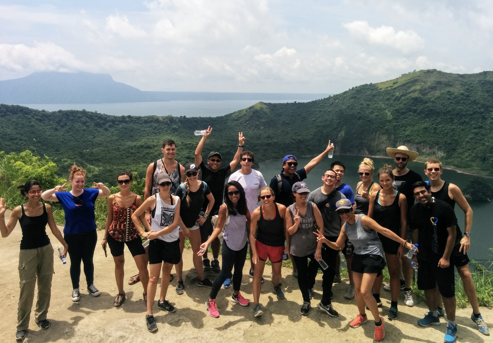 A group of 20 people on the peak of a volcano on a sunny day, with greenery in the background. They are all wearing various workout clothes, smiling at the camera, and some are holding up peace signs.