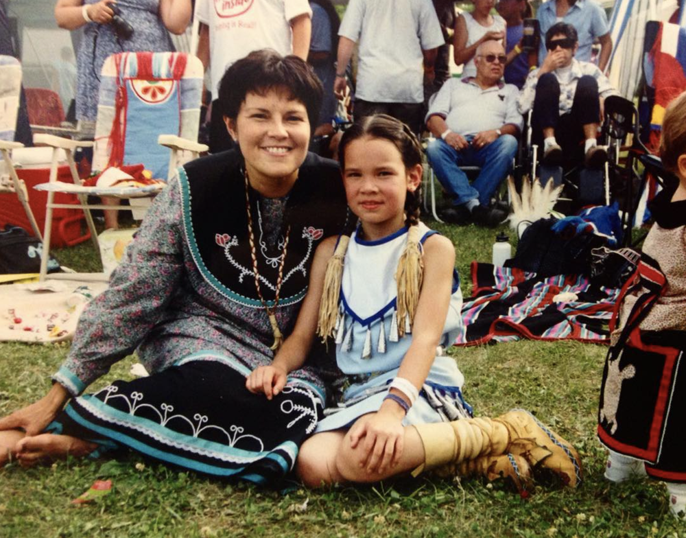 A mother and daughter sit on the grass with a crowd behind them.