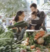 Im Vordergrund Gemüse auf einem Markt, dahinter Suelyn mit ihrem Mann, der ein kleines Kind vor sich in einer Tasche trägt