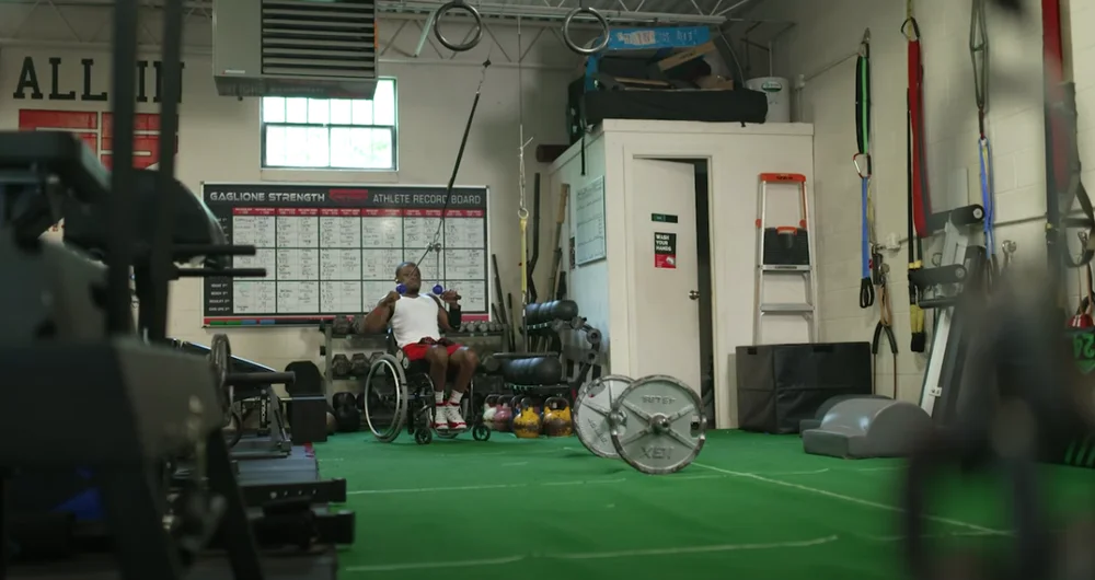 Garrison Redd, a male individual who uses a wheelchair, pulls a strength band from the ceiling of a gym. He is surrounded by various athletic weights and equipment.