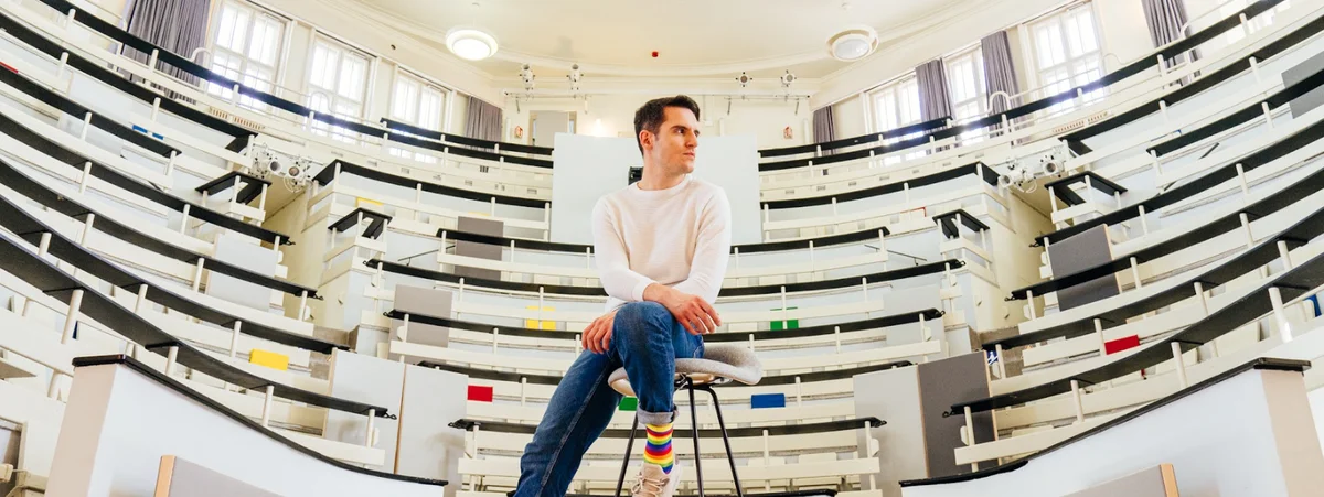 Emre Celik sitzt auf einem Barhocker und schaut rechts an der Kamera vorbei. Hinter ihm ist das Auditorium im Eventbereich des Berliner Google-Büros zu sehen.