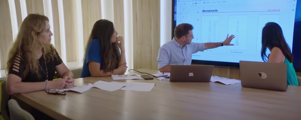 Four people sitting at a conference table. Two of them have laptops open. They all swiveled in their seats looking at sketch work on a large screen behind them. One person gestures toward it.