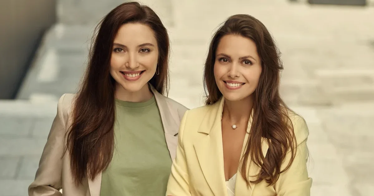 Two women with brown hair smile at the camera in front of a flight of stairs. One is wearing a green shirt and cream blazer, and the other a yellow blazer.