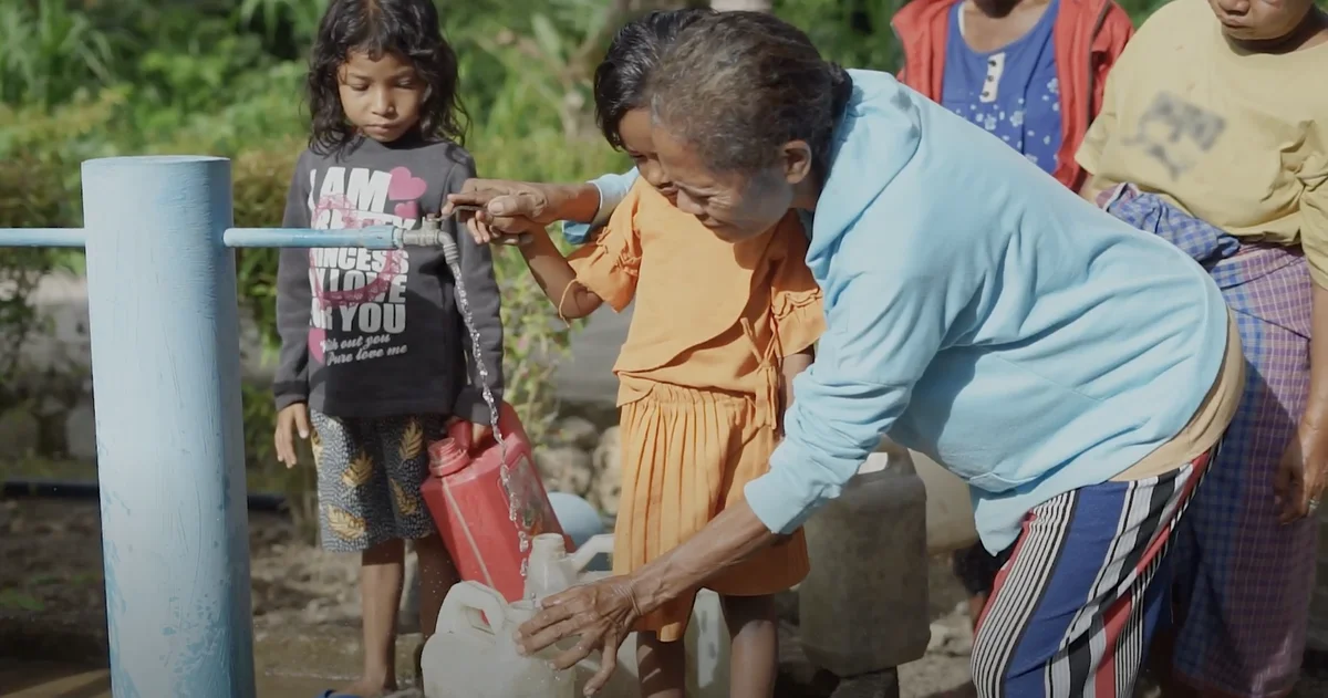 A group of women and children stand outside by a water faucet, pouring water into a container.