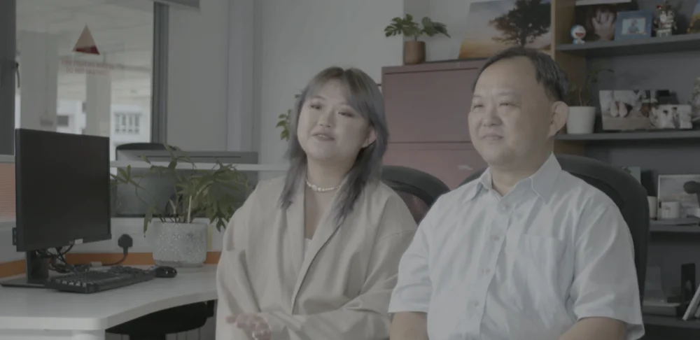 Two people sitting down by a desk with a computer, with a busy shelf behind them.