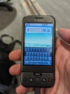 An Android G1 smartphone in someone’s right hand with the virtual on-screen keyboard visible. The digital keyboard is pulled up revealing all lowercase letters.