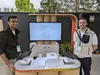 Two Googlers stand in front of laptops, with a large TV screen behind them showing a Gemini Advanced conversation. On the table between the laptops are a printed out economics textbook and a huge stack of 1,500 pages of paper.