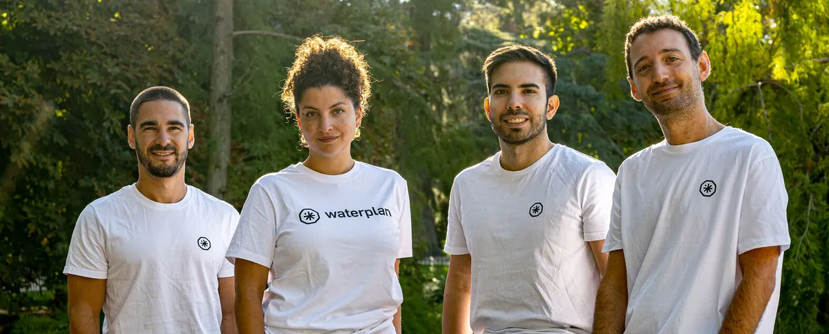 Four people wearing white t-shirts with blue writing that reads "Waterplan" look at the camera outdoors. Behind them is a sunny grove of trees.