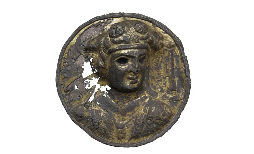 A rusty bronze coin with bronze still showing in some parts, with a relief of a face looking at an angle,  with hollowed eyes.