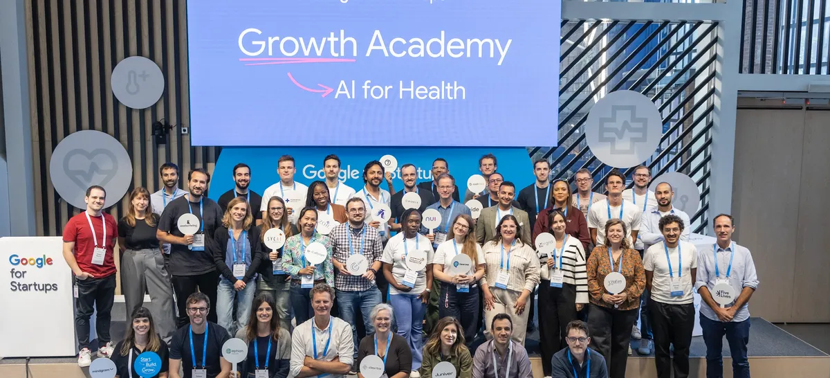 A group of people stand in an auditorium, looking at the camera and holding up signs with their company logos, in front a blue screen that reads "Growth Academy: AI for Health"