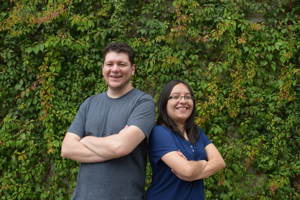 Two people stand back-to-back with their arms crossed. One is wearing a gray t-shirt and the other is in a blue t-shirt standing in front of a plant-covered wall.