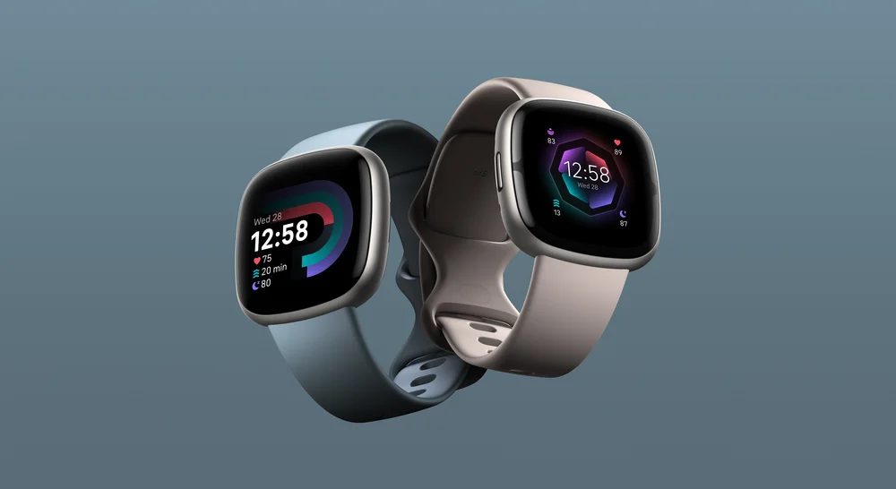 Versa 4 in waterfall blue and Sense 2 in lunar white photographed  side-by-side, showing a clock face with heart rate, Sleep Score and Active Zone Minutes.