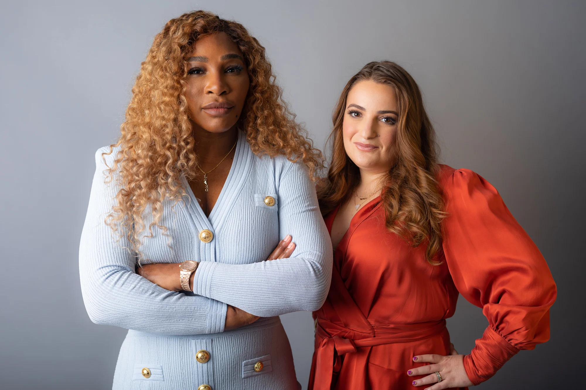 Serena Williams folds her arms and wears a light blue sweater with bronze buttons. Alison Rapaport Stillman places her arms on her hips and wears a bright red dress with long sleeves and a matching red belt.