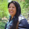 A woman with long dark hair and turquoise jewelry is pictured smiling in front of greenery.