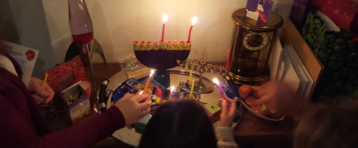A woman, two children, and a man holding candles facing a menorah that already has two candles lit and placed upon it.