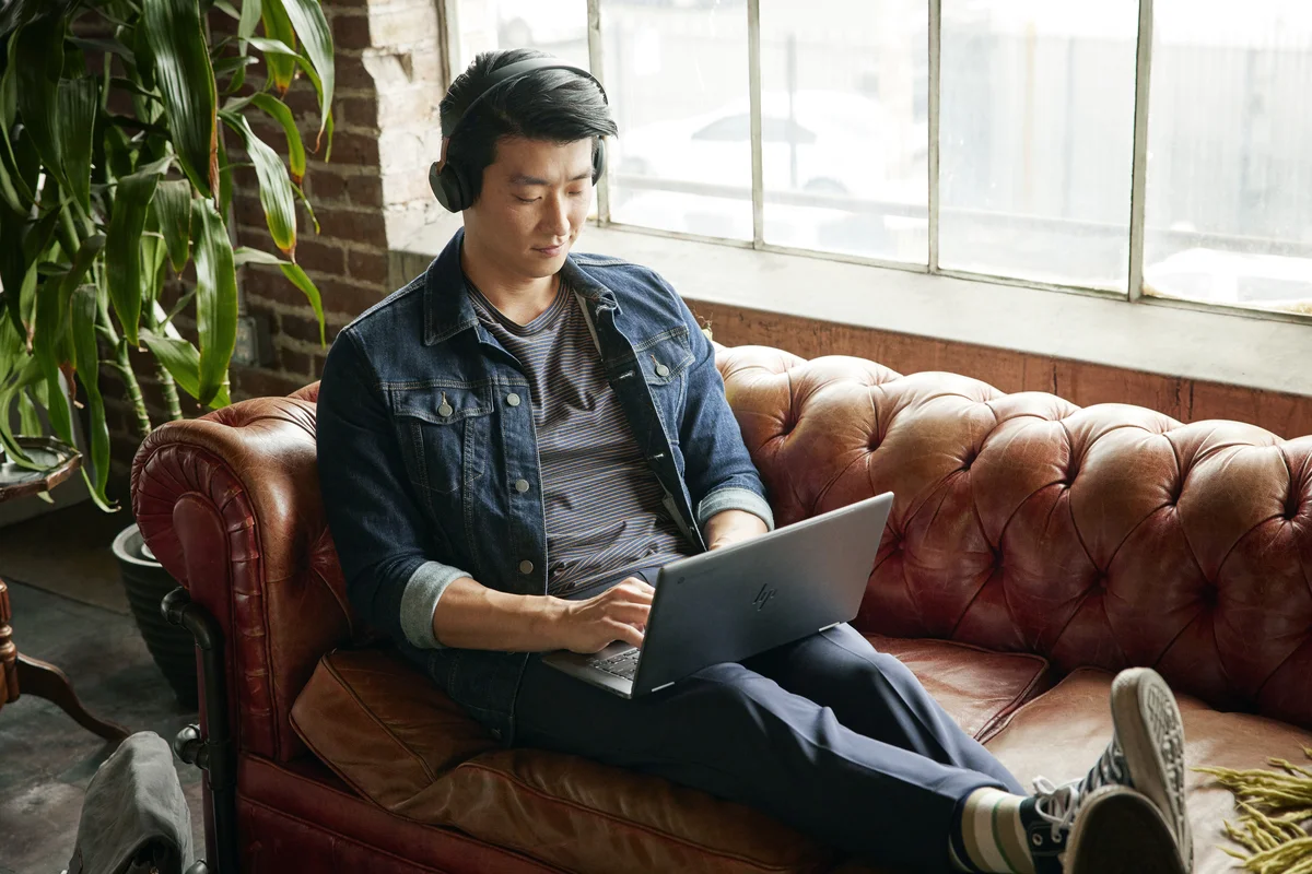A man wearing over ear headphones sits cross legged on a brown leather couch, while working on a Chromebook laptop in his lap.