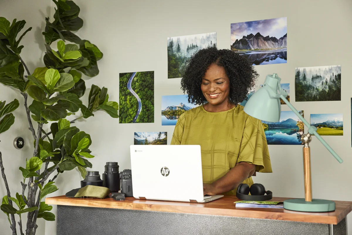 A woman in a yellow shirt stands at her desk, while working on an HP Chromebook laptop.