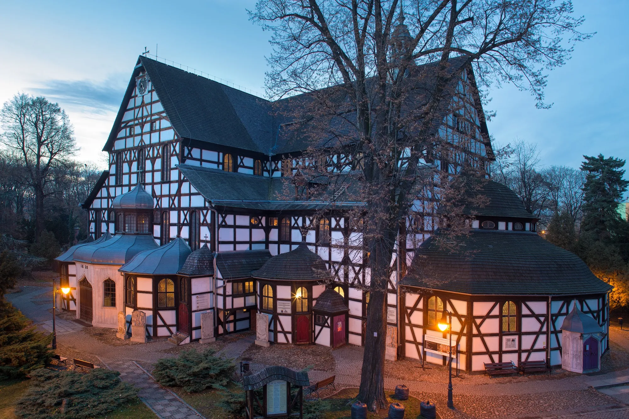 In the photograph there is a wooden church. The church is mostly white, the wooden elements are made of dark, timbre wood. The photo was taken after dusk - the sky is of navy blue colour. The street lamps emit a dim light.