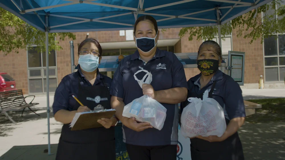 Three women in face masks and hair nets holding bags with food in them.