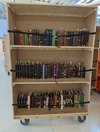 A wooden bookcase on wheels with three shelves and black straps around each shelf row.